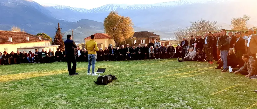 Residents gathered outdoors in a village square for a community consultation meeting, mountains behind